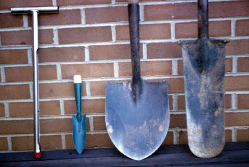 From left to right: a stainless steel soil probe that is a slim metal tool with sturdy grip at the top, and it looks like "T" shape, a hand garden trowel that has short handle and has a curved scoop with sharp top, shovel which has long handle and flat curved head , and a spade which has a flat, square-shaped head and short handle..