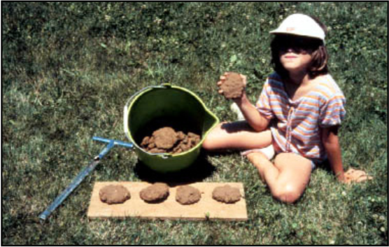 A girl sitting with a mud pie in her hand and she has more mud pies on a piece of wood.