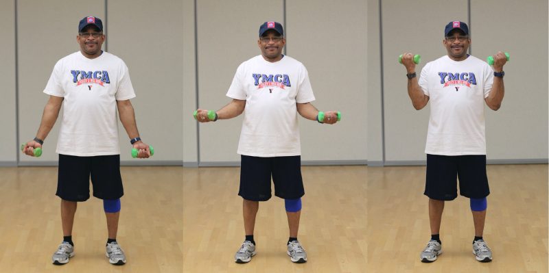 Images showing a man standing with his feet at hip-distance apart and his hands down by his side holding dumbells facing out. He bends at his elbows to bring his fists close to his shoulders and his elbows are close to his sides.