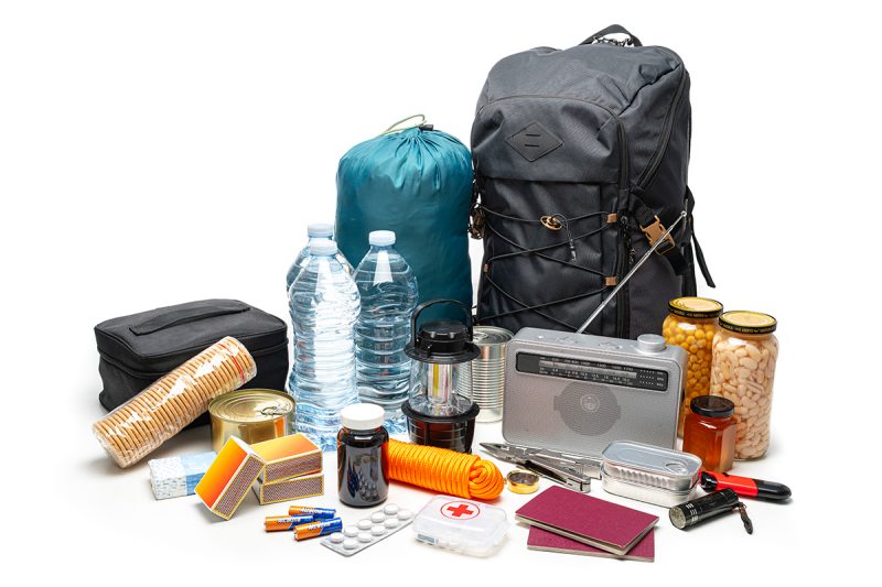 A collection of emergency preparedness items. There is a large black backpack, a teal sleeping bag, two large bottles of water, a black lantern, a silver radio with an antenna, two jars of preserved food (one with chickpeas and the other with white beans), canned food items, a roll of crackers in plastic packaging, several boxes of matches, batteries, medication bottles and blister packs of pills, an orange rope, a first aid kit with a red cross symbol on it, passports or notebooks with maroon covers, and various tools including pliers and scissors.