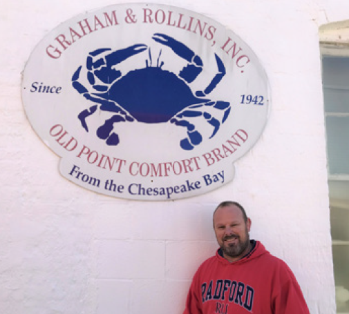 photo of Casey wearing a red hoodie, standing Infront of the logo of Graham and Rollins INC which is a huge blue crab with the name of the company