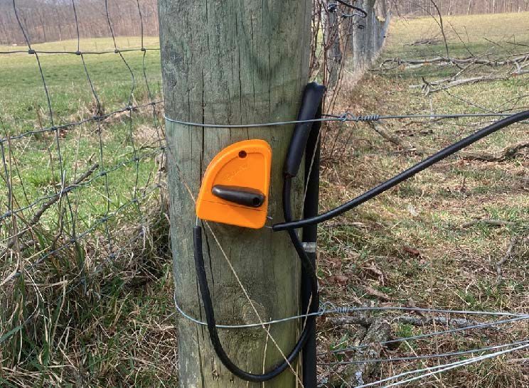 An orange Gallagher cut-out switch with a black on/off handle attached to a fence post, with two insulated cables extending out from the bottom on either end.